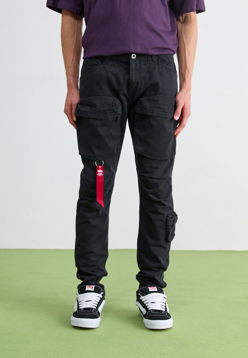 Black cargo pants with multiple pockets, a fabric loop with a red accent, and a relaxed fit, paired with black and white sneakers.