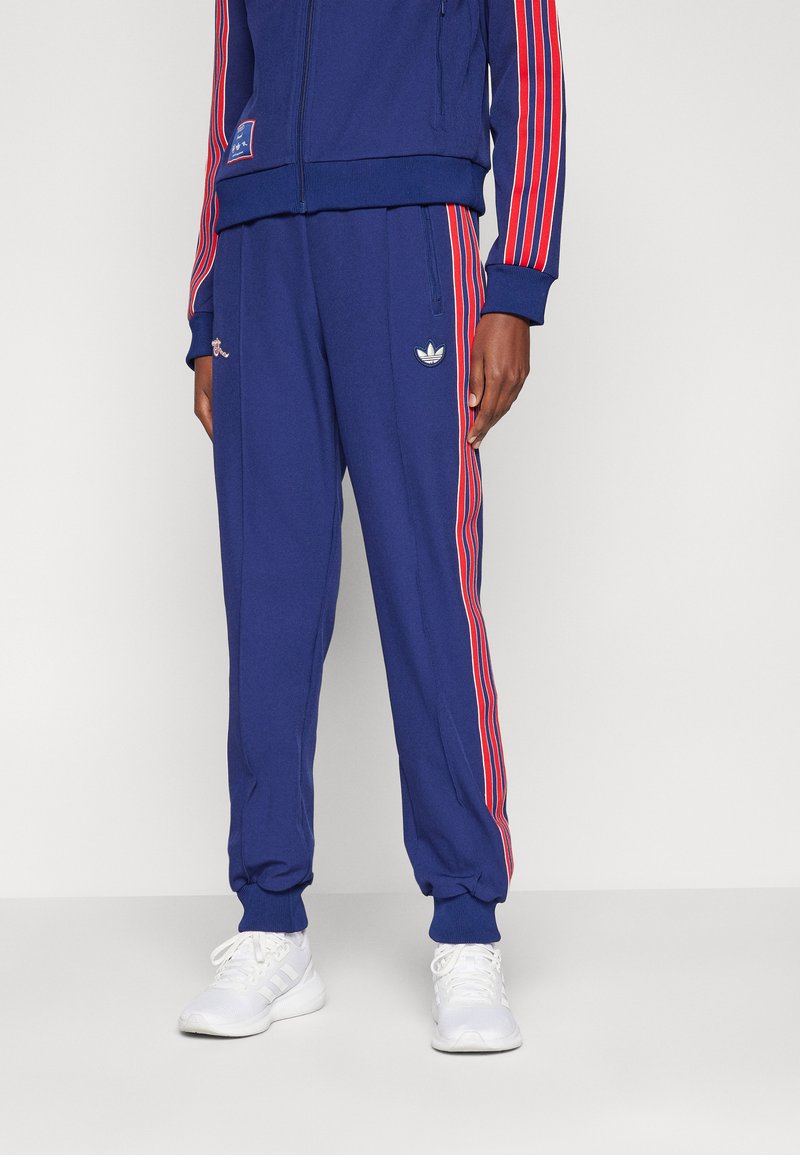 Blauer Trainingsanzug mit roten Streifen, Jacke mit Reißverschluss und Hose mit seitlichen Taschen und Logo-Akzenten. Hergestellt aus einem weichen, strukturierten Stoff.