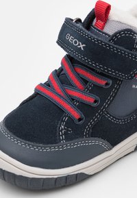 Zapato tipo bota alta en ante azul marino, con cordones rojos y una tira de Velcro. Detalles en cuero liso y suela de goma con textura antideslizante.