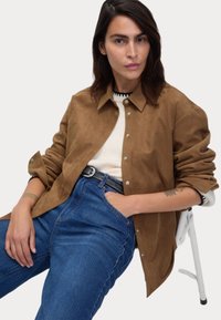 Chemise en daim marron avec fermeture à boutons, superposée à un pull crème. Jean en denim avec une ceinture noire, assis sur une chaise blanche.