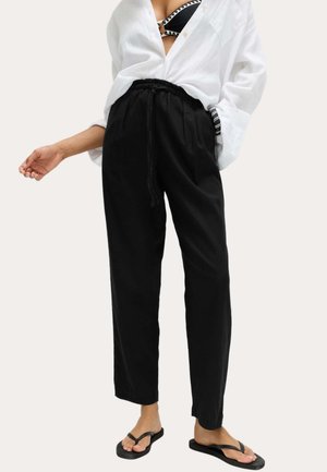 Vrouw draagt zwarte broek met trekkoord, half open wit overhemd waar een zwarte bikini top met patroon onder zichtbaar is, en zwarte slippers.