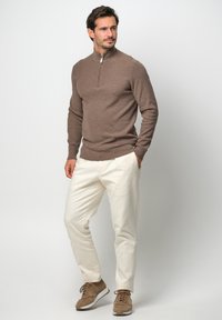 Brauner Pulli mit Reißverschluss am Hals, gefertigt aus strukturiertem Stoff, kombiniert mit cremefarbener, tapered Hose und braunen Sportschuhen. Schlichtes, modernes Design.
