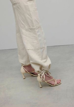 Pantalons cargo en lin crème avec poches, associés à des sandales à talons hauts nud avec lanières, présentant un bout carré et un talon évasé.