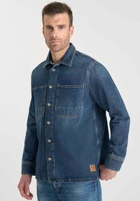 Blauwe denim jas met twee borstzakken, sluiting met knopen aan de voorkant en een gestructureerde afwerking. Bevat een logo patch aan de onderkant links.