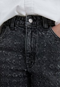 Jeans negros con un patrón floral texturizado, que cuentan con un cierre de botón y un acento de calavera plateada, junto a una camisa blanca.