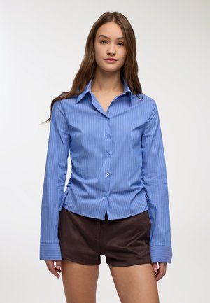 Giovane donna che indossa una camicia a righe blu con bottoni e maniche lunghe e pantaloni corti marrone scuro, in piedi davanti a uno sfondo bianco semplice.