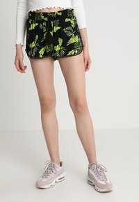 Pantaloncini neri con motivo di foglie verdi, vita elastica con cordoncino, e pizzo lungo l'orlo, abbinati a sneaker rosa chiaro.