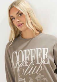 Sudadera gris con la frase "COFFEE Club" impresa en blanco, de corte relajado y textura suave. El diseño incluye ilustraciones relacionadas con el café.