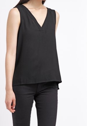 Top sin mangas negro con un escote en V profundo, hecho de una tela suave, que presenta un corte relajado y un dobladillo trasero ligeramente más largo. Combinado con pantalones oscuros.