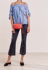 Blusa azzurro chiaro off-the-shoulder con dettagli a fiocco, abbinata a jeans scuri strappati e mule nere. Una piccola borsa a tracolla rossa in pelle completa il look.