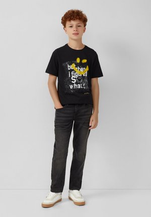 T-shirt nera in cotone con testo grafico bianco e giallo e faccina sorridente, abbinata a jeans scuri e sneakers bianche con suole beige.