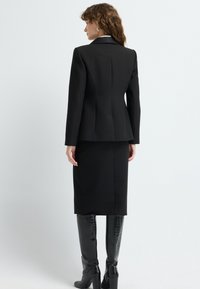 Černý přizpůsobený blazer s těsným střihem, spárovaný s černou sukní ke kolenům. Lesklé černé kozačky až po kolena dotvářejí outfit.