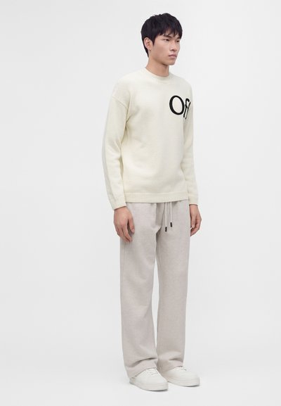 Homme portant un pull crème avec des lettres noires, un pantalon gris clair à cordon et des baskets blanches, debout devant un fond blanc uni.