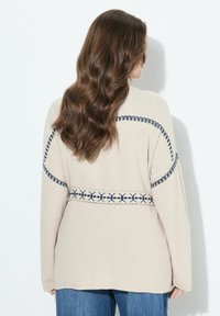 Beige stickad tröja med marinblå sömmar, med ett mönstrat band runt midjan och axlarna. Lätt passform och textil med struktur.