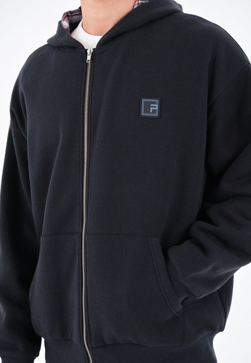 Schwarze Zip-Up-Hoodie mit Karolining, ausgestattet mit einer Fronttasche und einem gestickten Logo auf der Brust. Weicher Stoff mit lässiger Passform.