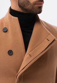Cappotto doppiopetto tan realizzato in tessuto morbido, con bottoni neri, colletto alto e un design pulito con dettagli di cucitura minimi.