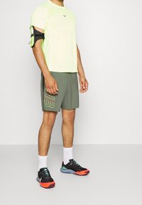 Neonově zelené atletické tričko s krátkým rukávem a geometrickou texturou, doplněné olivově zelenými šortkami s výrazným logem. Černé a červené atletické boty.