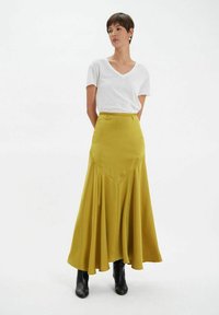 Une longue jupe jaune en tissu lisse, avec un design évasé et un détail de couture en V, assortie à un chemisier blanc à col en V et des bottes noires.