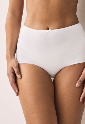 Środkowa część kobiecego ciała ubrana w białą bieliznę z wysokim stanem z małą kokardką, stojąca na neutralnym tle, z rękami spoczywającymi wzdłuż boków.