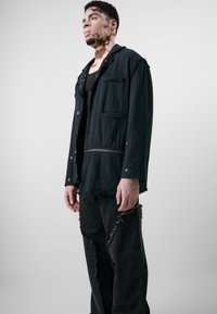 Veste noire avec un ourlet effiloché, présentant une poche zippée et des fermetures à bouton. Assortie à un pantalon noir usé montrant des bords effilochés.