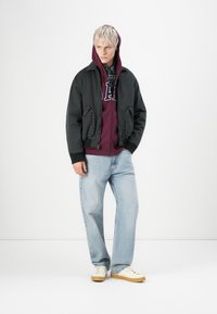 Zwarte bomberjack over een bordeauxrode hoodie, lichtblauwe jeans en beige sneakers. Heeft ritssluitingen en een gelaagde capuchon.