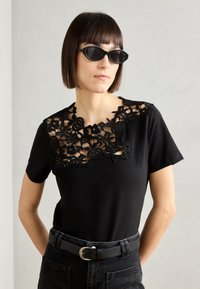 Sort t-shirt med blonder i halsudskæringen, korte ærmer og en tætsiddende pasform. Kombinationen med sorte højtaljede jeans og et bælte.
