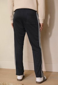 Pantalones de chándal azul marino hechos de un material suave, con rayas laterales blancas y un bolsillo en la parte trasera. Combinados con zapatillas blancas sobre un suelo de madera.