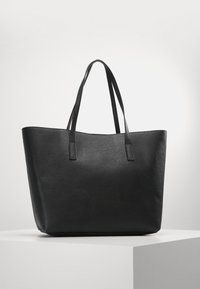 Sac fourre-tout en cuir noir avec une finition texturée, doté de deux longues poignées et d'un design à ouverture supérieure, posé sur un piédestal blanc.