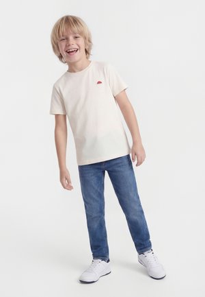 Smilende blond dreng iført en cremefarvet t-shirt, blå jeans og hvide sneakers, stående på en hvid baggrund.
