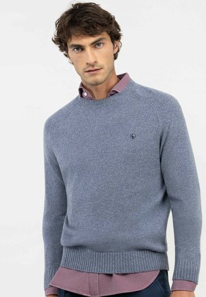 Hombre con suéter de punto gris claro sobre camisa con botones color malva y pantalones oscuros sobre un fondo blanco liso.