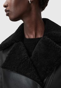 Czarna kurtka z owczej wełny, z dużym kołnierzem, teksturowanym materiałem i gładkim skórzanym wykończeniem. Projekt podkreśla ciepło i komfort.