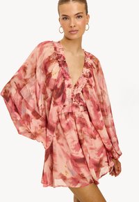 Vrouw in een vloeiende, roze en rode bloemenprintjurk met lange, luchtige mouwen en een diepe V-hals, gecombineerd met gouden ringoorbellen.