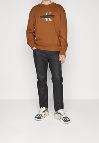 Sudadera marrón con el logo "CK", jeans negros y zapatillas beige/verdes. Ajuste casual, con puños acanalados y detalle en el dobladillo de la sudadera.