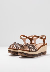 Sandalias de cuña de ante marrón con correas decorativas trenzadas en patrones de serpiente; textura trenzada en la cuña y diseño de punta redonda.