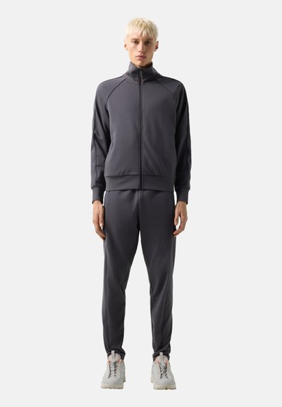 Jong persoon met kort blond haar staat gekleed in een donkergrijze jas met ritssluiting, bijpassende trainingsbroek en lichtgrijze sneakers tegen een witte achtergrond.