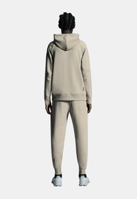 Beige hoodie en jogger-set gemaakt van zachte stof, met een ontspannen pasvorm, geribbelde manchetten en logo-accenten op de mouw en het broekspijpen.