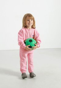 Felpa rosa in pile e pantaloni abbinati indossati da un bambino, che tiene un pallone da calcio verde e nero, in piedi su un pavimento liscio e chiaro.