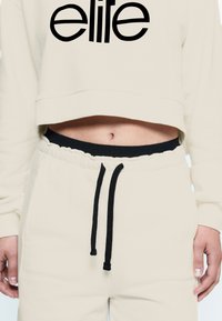 Sweatshirt corto crema con il testo "elite" in nero, abbinato a joggers coordinati con elastico in vita e coulisse nera.