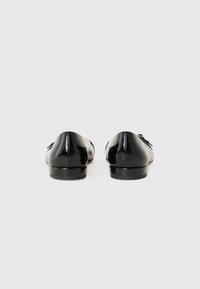 Mocassins en cuir verni noir avec une texture lisse et un talon bas, présentant un accent de nœud décoratif à l'avant.