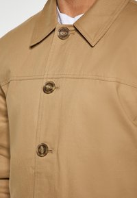 Beige knapjakke med skjortekrave, enkelt brystlomme og fire strukturerede knapper. Stoffet ser glat og holdbart ud.