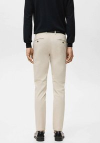 Pantaloni beige a taglio slim, con due tasche posteriori chiuse da bottoni, abbinati a un top nero a maniche lunghe e scarpe nere.