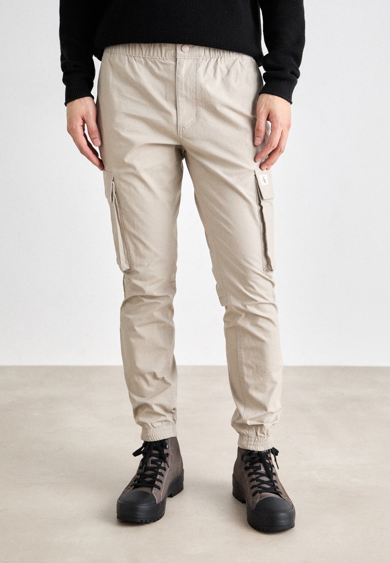Pantalones cargo beige con cinturilla elástica, bolsillos laterales con cremallera y piernas ajustadas. Combinados con botas oscuras de diseño robusto.