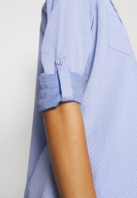 Rolled-up sleeve of a light blue patterned shirt with a buttoned tab securing the cuff against a neutral background.