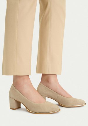 Kobieta nosząca beżowe zamszowe buty na klockowym obcasie ze złotą obwódką, w połączeniu z beżowymi skróconymi spodniami, stojąca na białym tle.