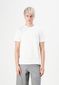 Weißes Baumwoll-T-Shirt mit Rundhalsausschnitt und kurzen Ärmeln, mit einem kleinen grauen Logo auf der linken Brust; getragen mit grauen Shorts.