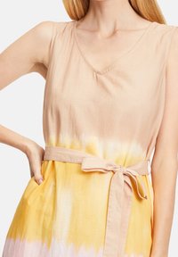 Šaty s tkaným vzorem v odstínech tan a žluté, s výstřihem do V, bez rukávů, látkovým páskem v pase a texturovaným, lehkým materiálem.