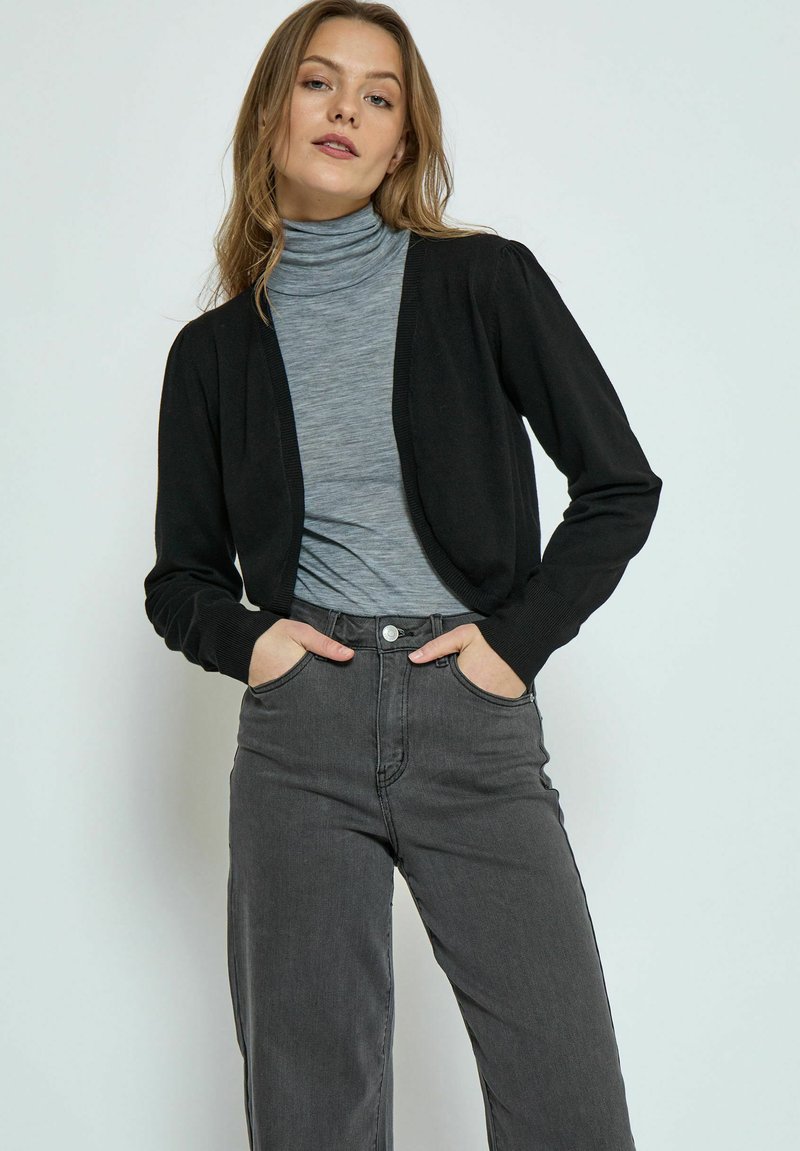 Schwarze, verkürzte Strickjacke mit V-Ausschnitt, langen Ärmeln und gerippten Bündchen, getragen über einem grauen Rollkragenpullover und hoch taillierten grauen Jeans.