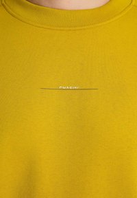 Sudadera amarilla con cuello redondo acanalado; presenta un pequeño texto de logo blanco "CHASIN'" con una línea horizontal debajo. Textura suave.