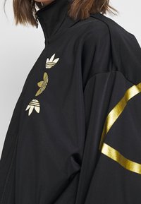 Black zip-up jacket made of fabric featuring gold Adidas logo and three stripes design on the chest and a diagonal gold accent on the sleeve.