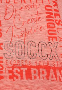 Camiseta roja de manga corta con una mezcla de texto impreso en negrita y adornos de pedrería. La tela tiene una apariencia texturizada y jaspeada.